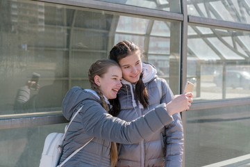 young teenager girls friend takes selfie by smartphone at city street on bus station with glass wall