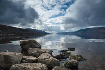 A view across Loch Ness looking down the length of the lake with rocks inn the foreground and dark clouds above, in Scotland, UK