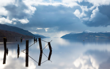 A view across Loch Ness with a broken pier in the foreground, and the length of the lake in the background and dark clouds above, in Scotland, UK