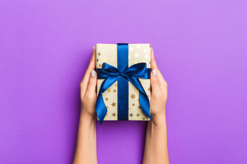 Flat lay of woman hands holding gift wrapped and decorated with bow on purple background with copy space. Christmas and holiday concept