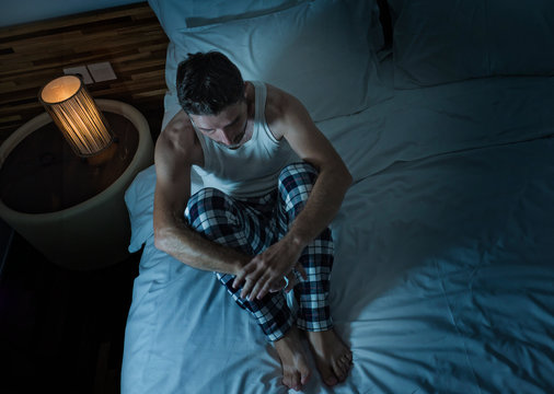 dramatic lifestyle portrait of young attractive sad and depressed man sitting on bed at night feeling stressed and desperate suffering depression problem in sadness and pain
