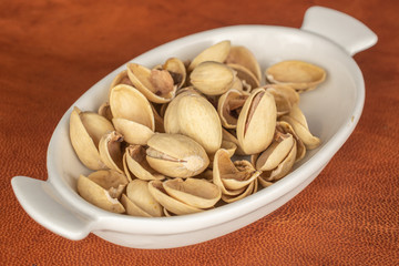 Lot of whole salted pistachio in small white ceramic bowl on cognac leather