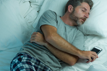 domestic lifestyle high angle portrait of young attractive and tired man sleeping on bed holding mobile phone in internet and social media addiction and network overuse