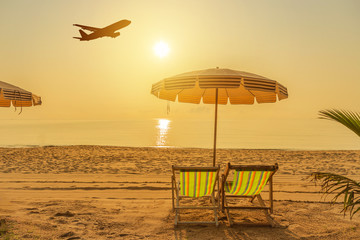 Empty wooden beach chairs and parasols on tropical sandy beach in the morning  with  silhouette airplane landing above the sea , destinations relax summer holidays concept 
