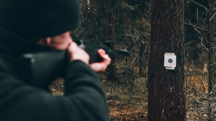 A man with a black air rifle in his hands shoots at a target fixed on a tree trunk in the forest,...