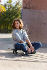 Girl teenager sitting on a skateboard. Beautiful fashionable skateboarder in jeans and a hoodie on a ramp.