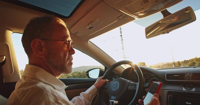 Stylish bearded business man using smartphone mobile driving car in front of beautiful sunlight rises on background. Communicate chat. Social networking. Adult traveling blogger lifestyle. Freelancer