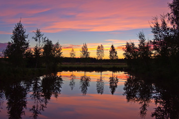 Beautiful colourful sky at dawn by a small pond, trees reflecting on calm water surface