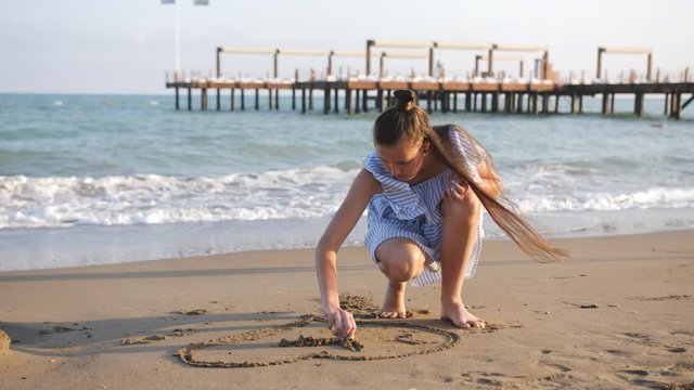 Girl teenager dress draws on the sand on the seashore.