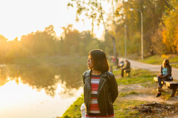 Fototapeta premium young brunette girl in a black jacket and striped t-shirt looks at the lake in the park