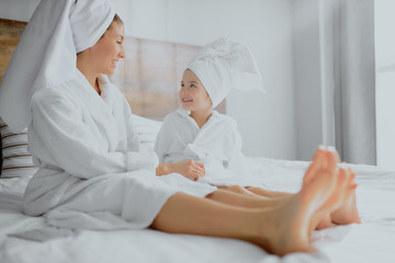 side view on lovely woman in white bathrobe and towel with cute daughter lying on bed in bright bedroom, little child look at mother