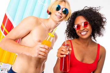 Pretty african female and caucasian blonde man stands in swimsuit with rubber beach mattresses, drinks juice and smiles, isolated on white background