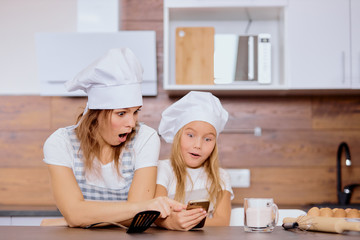 surprised woman and kid girl looking for recipe, before baking cakes, wearing aprons and caps in kitchen