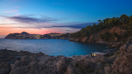 Fototapeta na wymiar Rocky bay with lonely hut and night city lights on the background of scenic sunset sky