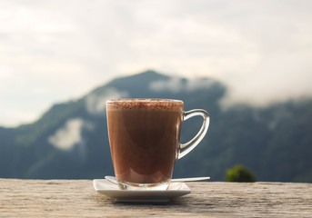 Hot chocolate in a glass cup on a wooden terrace with mountain view background in the morning light.