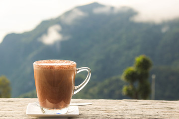 Hot chocolate in a glass cup on a wooden terrace with mountain view background in the morning light.