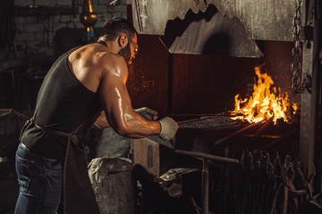 young caucasian strong man in black apron heating steel piece in furnace, powerful and strong...