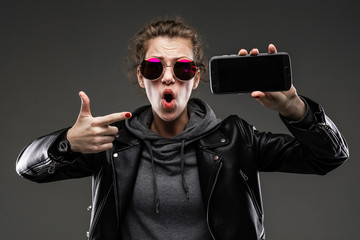 Stubborn caucasian girl with rough facial features in a black jacket shows her phone isolated on black background