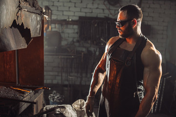 side view on strong forger in leather uniform and protective eyeglasses look at heating iron in furnace