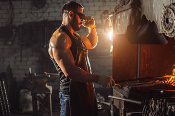 young strong muscular blacksmith wearing black apron and gloves for safety, stand preparing iron on...