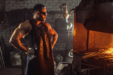 muscular adult with beard wearing leather apron working in workshop, heating. hardened metal in furnace