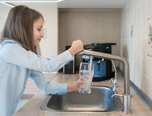 Portrait of a little caucasian girl gaining a glass of tap clean water. Kitchen faucet. Cute curly kid pouring fresh water from filter tap.  Indoors. Healthy life concept