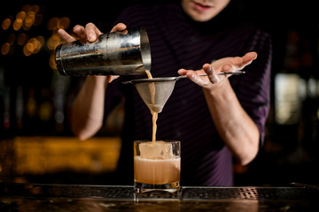 Fototapeta na wymiar Male bartender pouring an alcoholic creamy color drink from the steel shaker to the glass with ice cube through the sieve