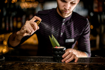 Male bartender spaying on a black glass with cocktail decorated with a tropical leaf, dried rose bud and essence drops