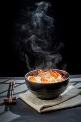 Noodles with steam and smoke in bowl on wooden background, selective focus. Asian meal on a table,...