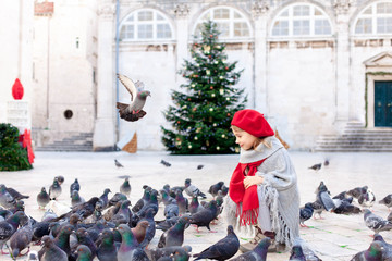 Christmas mood and holidays in Europe. Child girl feeds doves on square. Pigeons flying around cute...