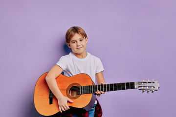 portrait of talented caucasian kid boy holding acoustic guitar and try to play on it, wearing casual clothes isolated over purple background