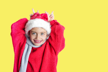 7 yaers old smiling girl with changing tees smiling in santa hat, red pullover, gray scarf, imitation of christmas dear with smile on yellow background. Natural light. Space for text