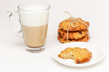 Transparent mug of hot latte and homemade whole grain crispy oatmeal cookies with raisins folded in a stack on white background. One cookie is bitten off. Concept of good start to the day. Closeup