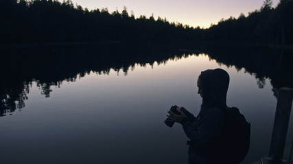 Silhouette traveler photographing scenic view in forest, river. Wood pier. One woman shooting nice dark magic night look. Girl take photo video on camera. Photographer walk with backpack. Outdoor.