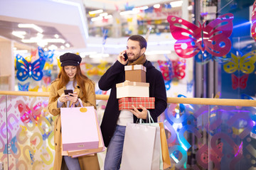 caucasian young couple with gifts and bags after shopping in shop center, redhaired woman and handsome bearded man together. man talking on phone, while his girlfriend look at screen of smartphone