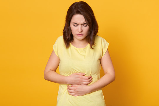 Frustrated brunette woman keeping hands on belly, feels pain as has menstruation or illness, wearing casual t shirt, stays at home, isolated over yellow background. Discomfort in stomach and period.