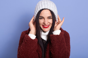 Beautiful adorable female wearing maroon faux fur coat, white sweater, winter cap, looks smiling with happy expression directly at camera, keeps hands on her head, isolated over blue background.