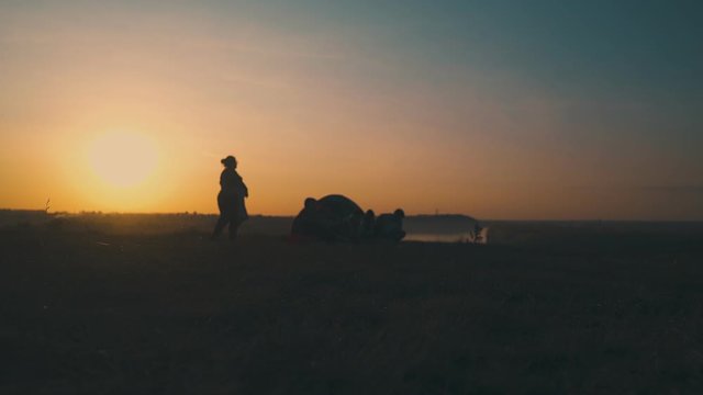 silhouette of overweight woman dancing near camping friends at bonfire on river bank against setting sun low angle shot