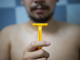 Selective focus on hand holding yellow Shaver with blur man has messy beard and mustache in bathroom background - 306732875