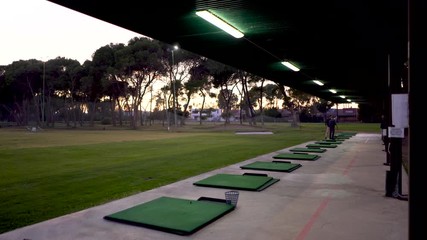 Golf monitor training golfer in practice area at sunset, golden sky with pine trees, green grass, in-line mats and both illuminated by artificial lights