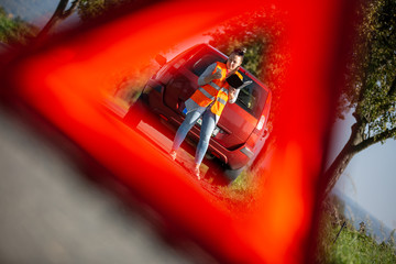Naklejka premium Annoyed young woman in a road distress situation - setting up a warning triangle and calling for assistance after her car broke down in the middle of nowhere; transportation concept (car problem)