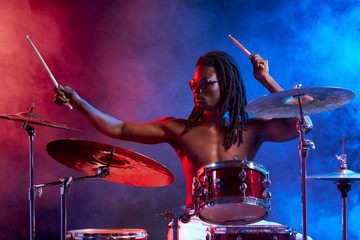 portrait of talented african man playing on drums, with naked skin wearing eyeglasses isolated over smoky neon background