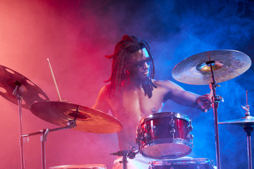 portrait of showy african drummer man with percussion instruments isolated over neon smoky...