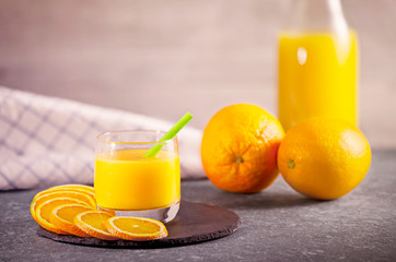 Delicious freshly squeezed orange juice in a small glass with fresh oranges, slices of orange and a bottle next to it on a rustic dark gray stone plate. Concept for healthy nutrition.