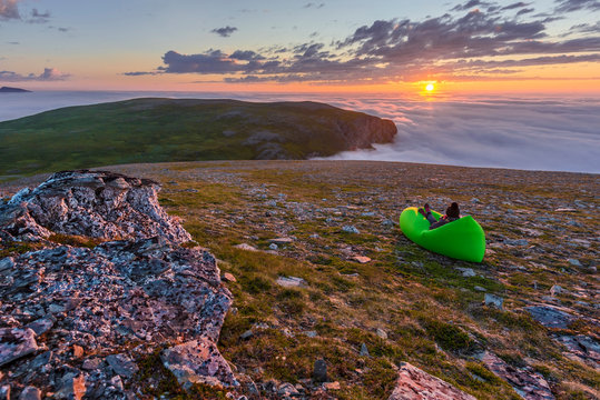 Woman in the air lounger is enjoying the midnight sun in the Northern cape of Soroya Island in Norwegian Finnmark. Fascinating sea of clouds is covering the Arctic Ocean surface at the background.