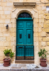 Fototapeta na wymiar Ornate wooden green door in a stone entry in Cospicua, Malta. With glass window above, iron fence below and potted plants on both sides. Architectural theme.