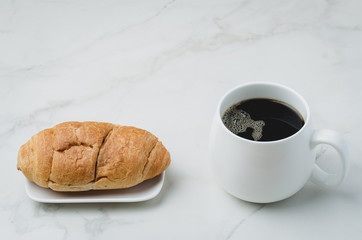 White mug with black coffee and croissant on white stone table. Coffee break.