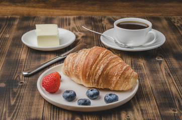 Coffee cup, croissant with berries in white bowl and butter knife on wooden table. Selective focus. Healthy breakfast with fresh berries