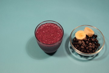 Blackberry and banana smoothie in glass, pot with berries and fruit slices on blue background