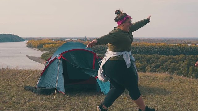 pudgy lady runs to girl friends and hugs by blue camp tent against calm river with forests on opposite bank slow motion